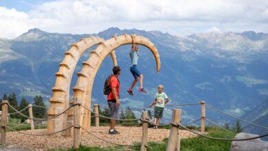Zwei hölzerne Steinbockhörner als Skultur ragen aus dem Boden. Kinder klettern darauf. Im Hintergrund sieht man Berge.