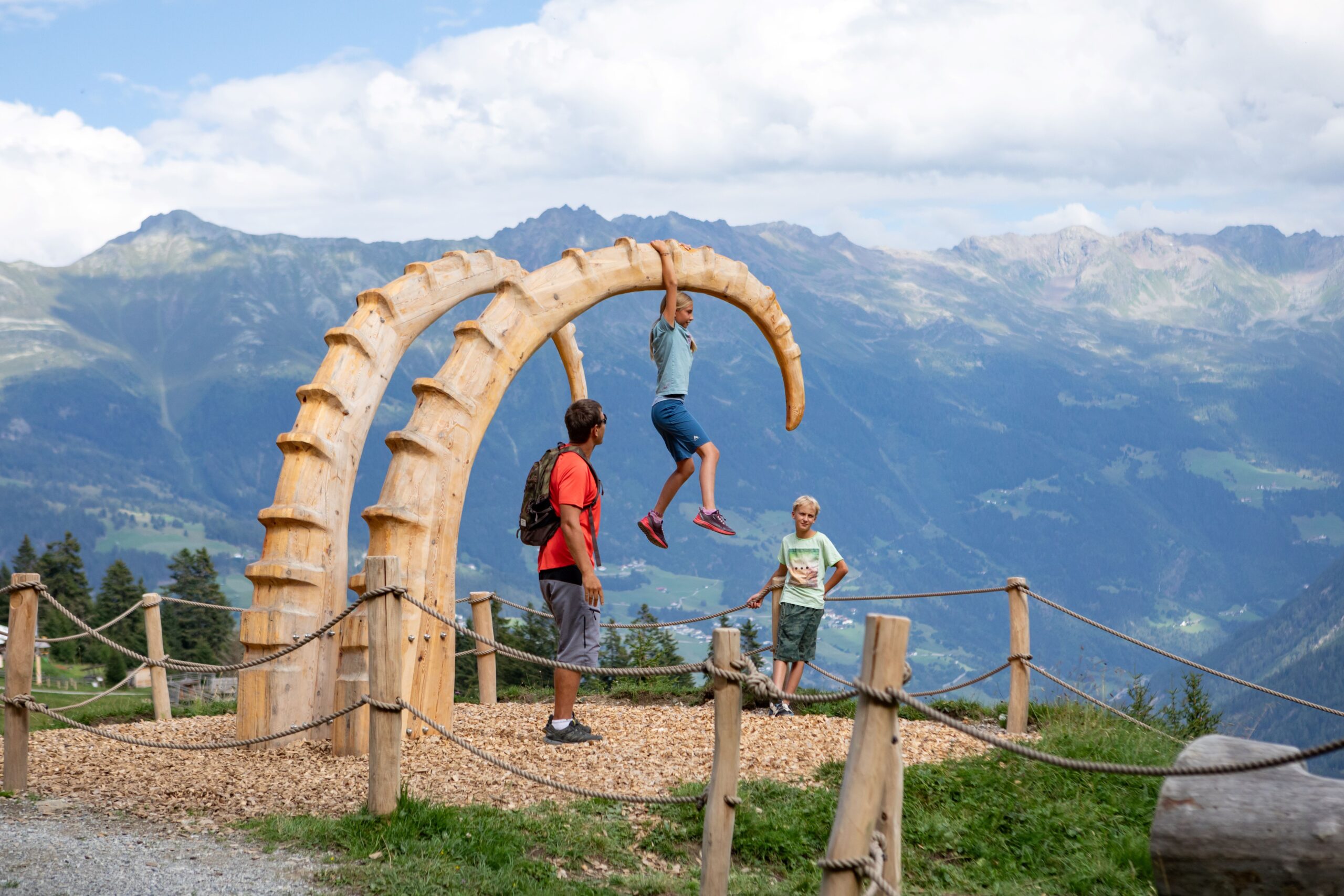 Zwei hölzerne Steinbockhörner als Skultur ragen aus dem Boden. Kinder klettern darauf. Im Hintergrund sieht man Berge.
