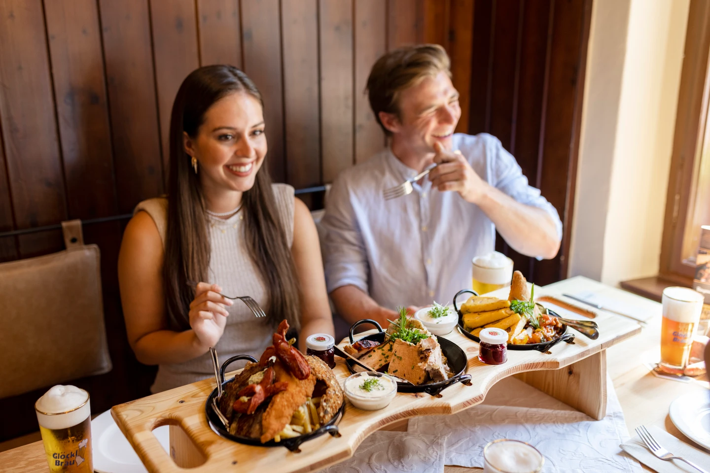 Zwei Menschen sitzen in einem Gasthaus und haben Teller mit Essen vor sich stehen. Sie wirken glücklich und lachen.