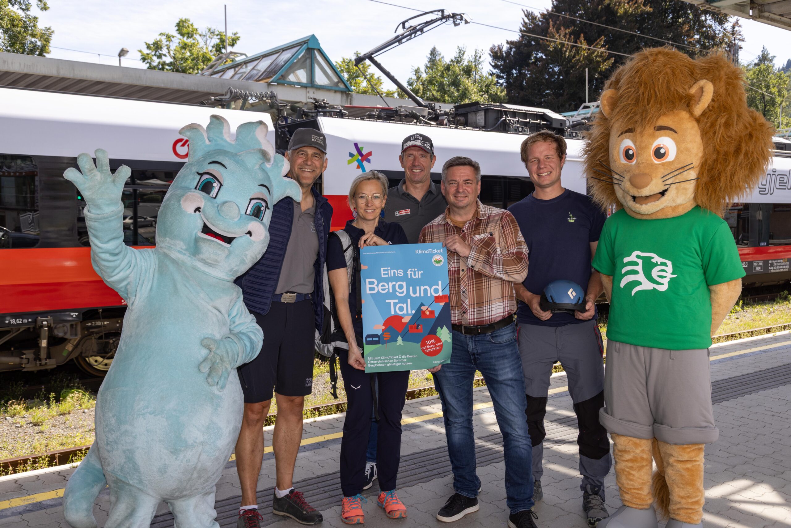 Ein Bild für eine Werbung zeigt fünf Personen zusammen mit zwei Maskottchen vor einer Bahn.