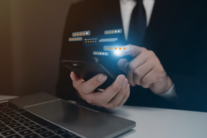 Ein Mann hält ein Handy in der Hand und zeigt auf einen Bildschirm mit einer Reihe von Sternen. Wie schafft man mehr Transparenz für Online-Bewertungen?